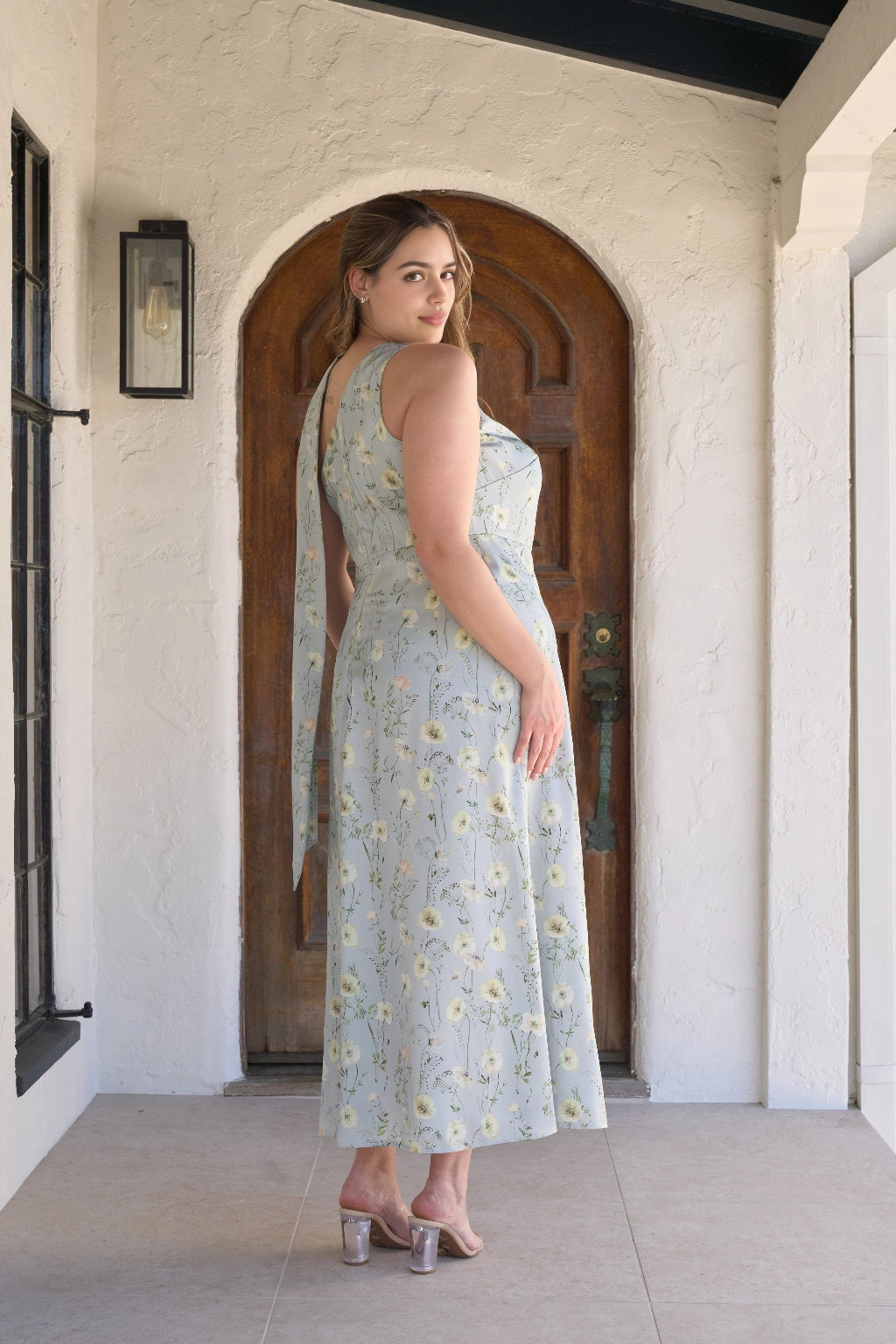 Back view of light blue floral plus size wedding guest dress featuring an asymmetric neckline, long draped sash and ankle-length skirt by roselledress