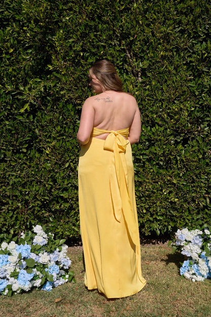 Back view of plus size yellow bridesmaid dress featuring open back, bow tie closure, long flowing ribbons and floor-length silhouette.