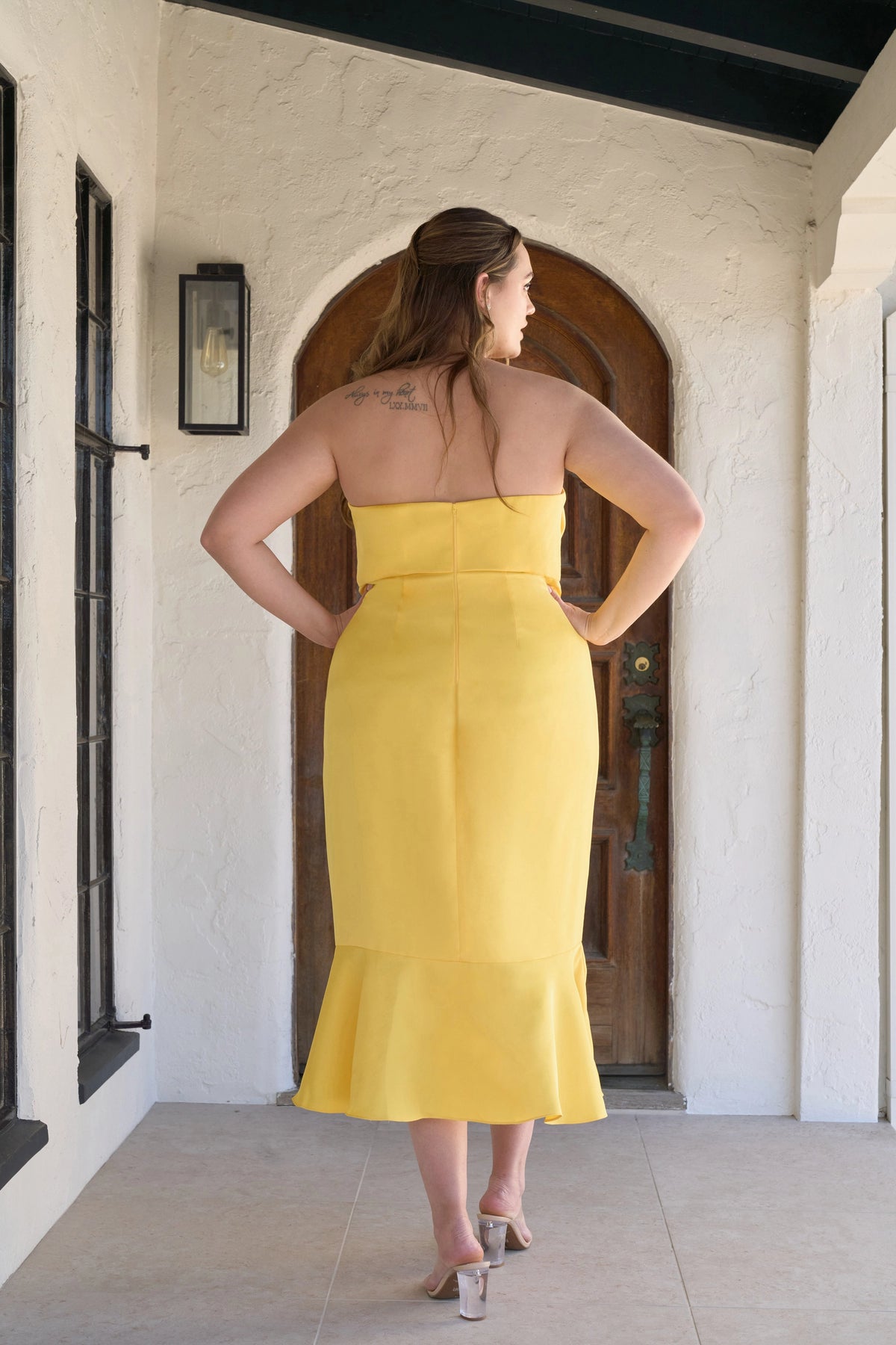 Back view of plus size yellow mermaid wedding guest dress featuring a straight strapless back, center zipper, fitted waist and flared midi skirt by roselledress.