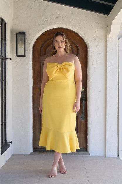 Plus size bright yellow strapless wedding guest dress with oversized bow neckline, fitted mermaid silhouette and flared tea-length hem.