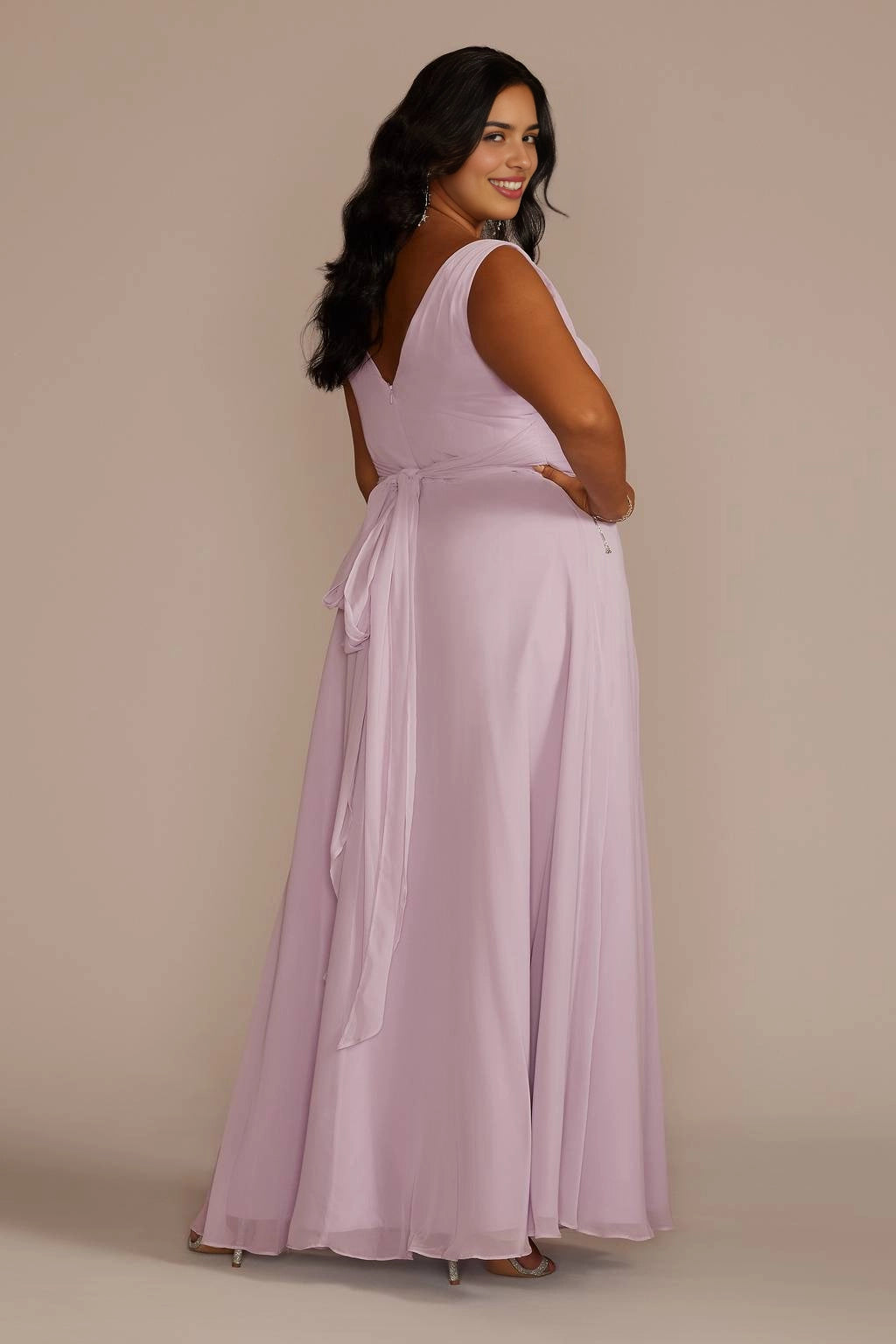 Back view of dusty lilac plus size wedding guest dress featuring a V-back, tied waist sash with flowing tails and floor-length skirt.